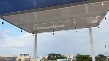 Ein überdachter Außenbereich mit Metalldach und hängenden Lichtern, mit Blick auf eine Straße mit Gebäuden und einer Tankstelle.