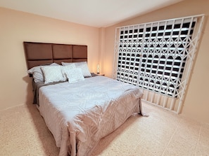 A bedroom with a bed, a headboard, a nightstand, a lamp, and a window with a metal grille. - Cozy 1-bedroom apartment in delightful Machala with AC (Machala)