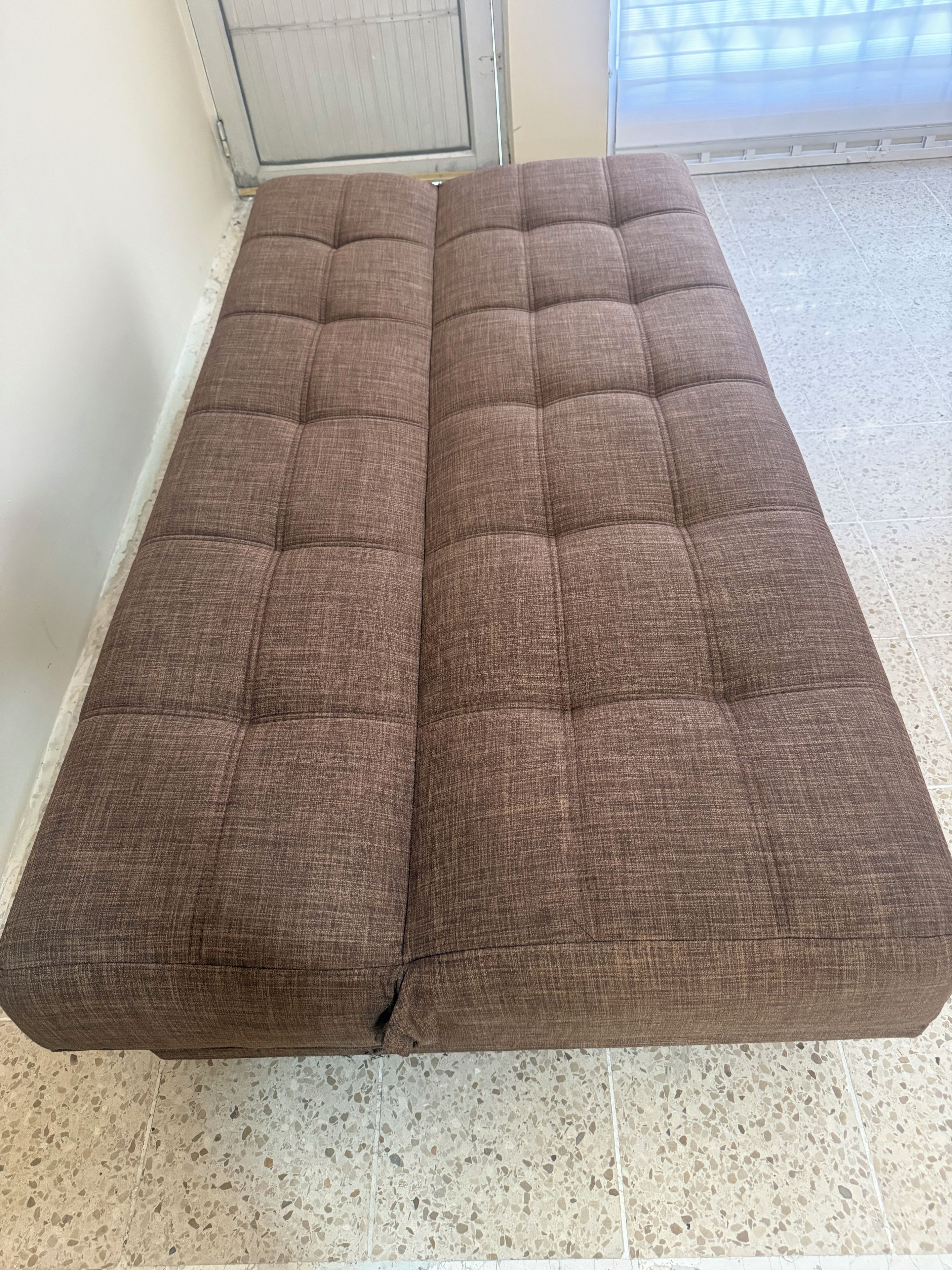 Un sofá marrón con diseño capitonado, ubicado en una habitación con piso de baldosas y una puerta al fondo.