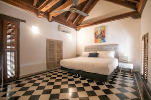 Quarto com piso xadrez, cama com cabeceira cinza, um quadro na parede e ventilador de teto.
