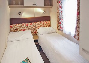A room with two single beds, a wooden headboard, a small table, and a window with patterned curtains. - New in 2017. 12ft-wide caravan with one double and two twin. (St. Leonards, Ringwood)
