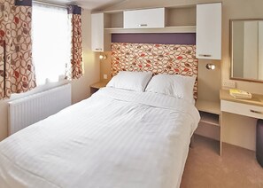 Een netjes opgemaakt bed met witte lakens in een kamer met een gestreept hoofdeinde, een nachtkastje met een lamp en een klein bureau met een spiegel en een telefoon.