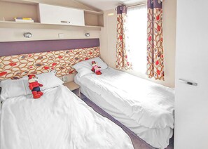A bedroom with two beds, a headboard with a patterned design, a shelf with books, and curtains with a geometric pattern. - 12ft-wide caravan with one double with en suite bathroom and one twin bedroom. (Milford-on-Sea, Nr Lymington)