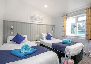 A hotel room with two beds, a window with curtains, a radiator, and a wall-mounted light fixture. - Select Lodge Plus 2 (Pet) (Milford-on-Sea, Nr Lymington)
