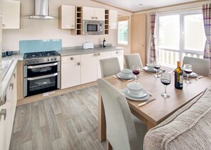 A modern kitchen with a gas stove, oven, microwave, and built-in cabinets. There is a dining table set with plates, glasses, and a bottle of wine. - Sublime 2 Caravan (Pet) (St. Leonards, Ringwood)