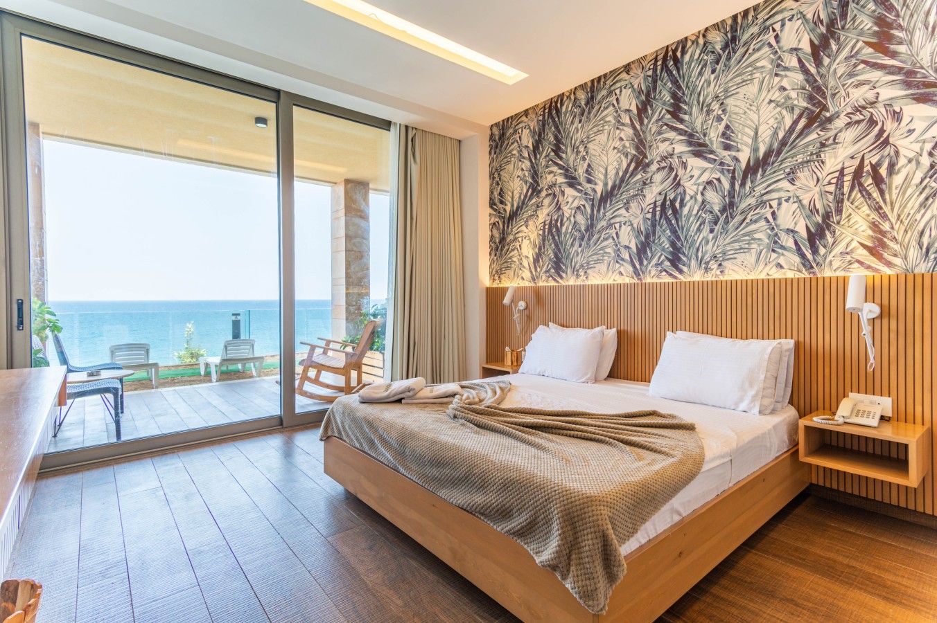 A hotel room with a large bed, a wooden headboard, a view of the sea, and a balcony with outdoor seating.