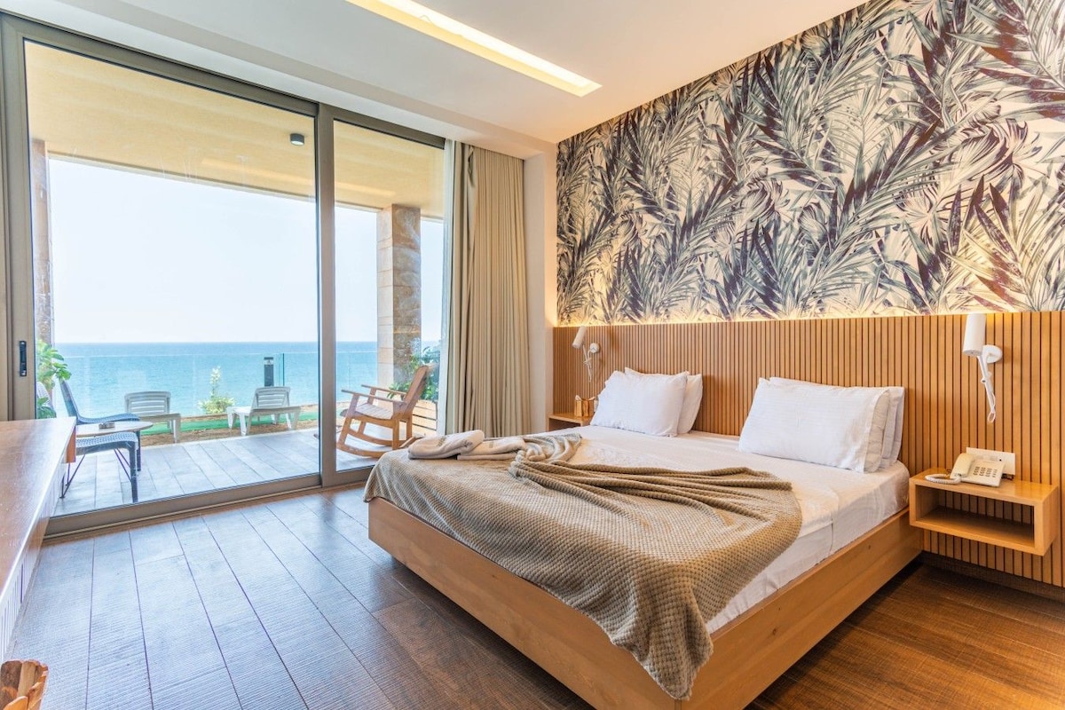 A hotel room with a large bed, a wooden headboard, a view of the sea, and a balcony with outdoor seating.