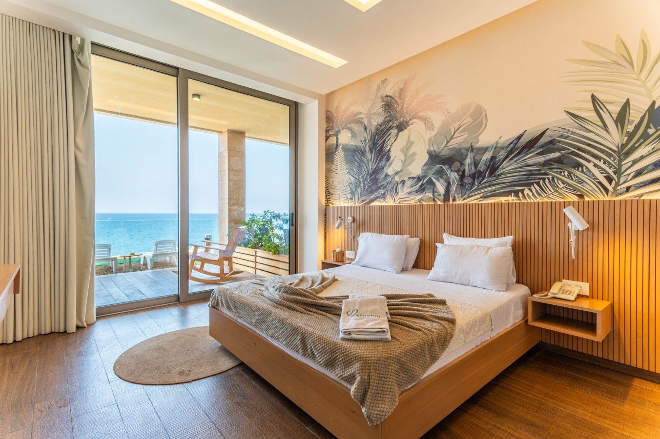 A hotel room with a large bed, a view of the sea, a wooden headboard, and a tropical mural on the wall.
