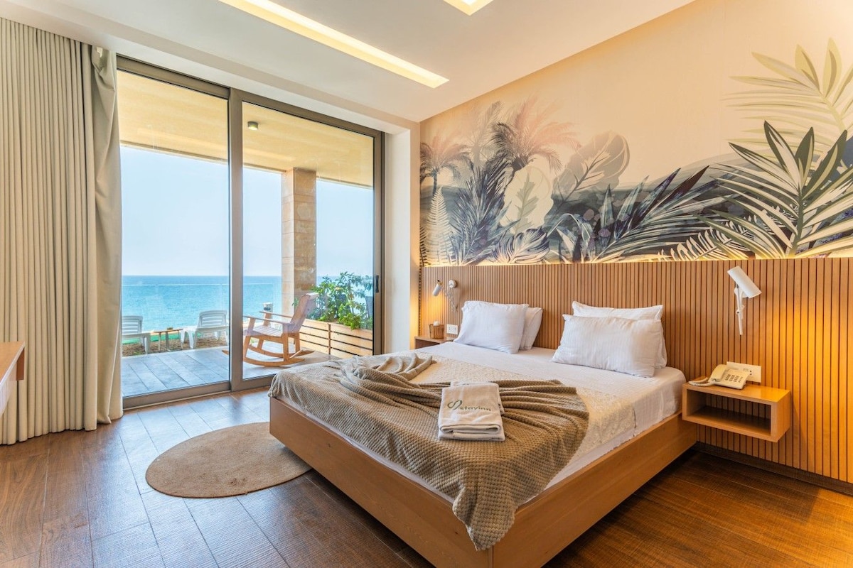 A hotel room with a large bed, a view of the sea, a wooden headboard, and a tropical mural on the wall.