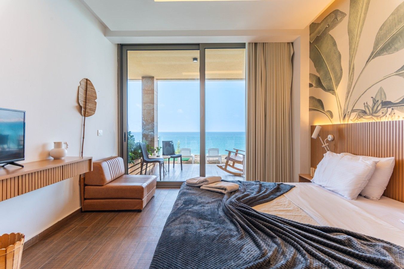 A hotel room with a bed, a TV, a seating area with a view of the sea, and a balcony with outdoor furniture.