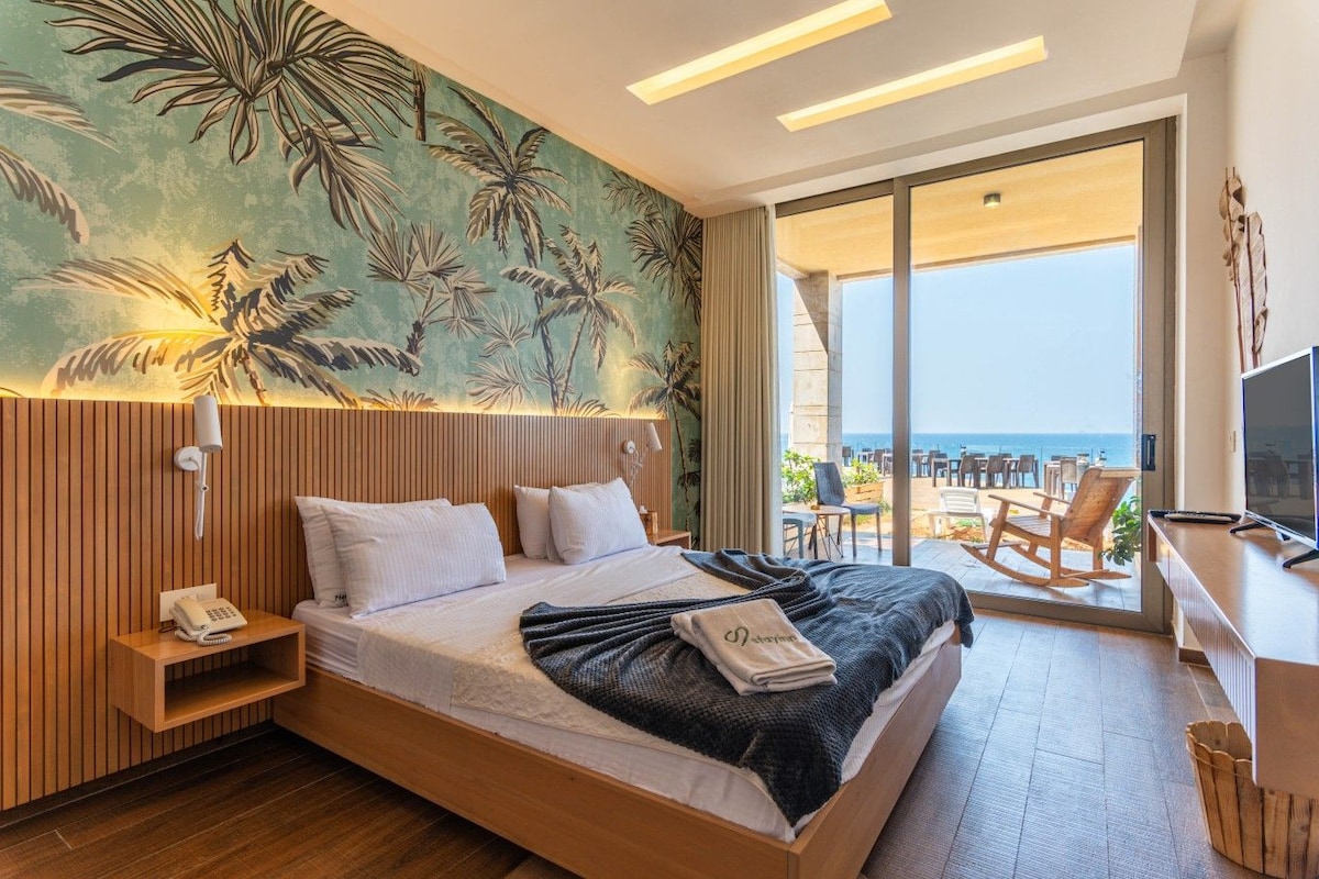 A hotel room with a bed, a desk with a TV, a phone, and a view of the sea.