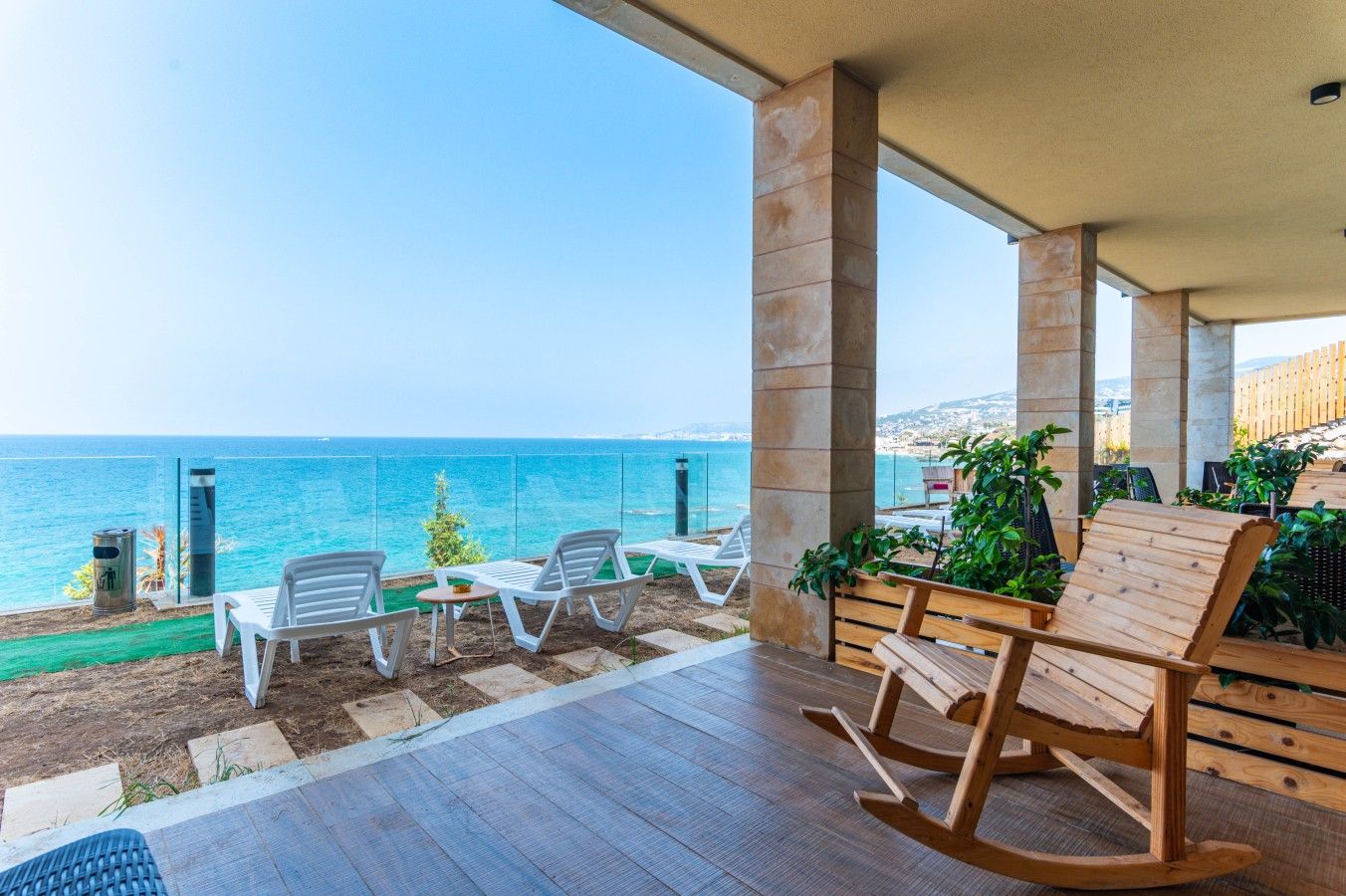 Un espace extérieur abrité, aménagé avec des sièges, offrant une vue sur la mer et comprenant un fauteuil à bascule en bois et des chaises longues blanches.