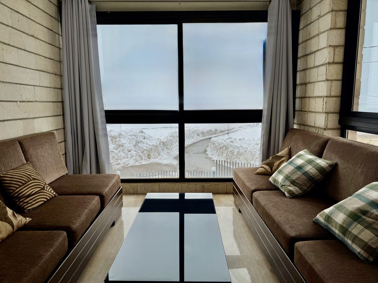 A modern living room with two brown sofas, a black coffee table, and a large window offering a view of a snowy landscape.