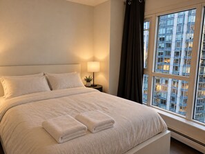 Quarto de hotel com cama grande, abajur, janela com cortinas e vista para um edifício.