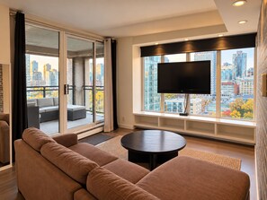 Uma sala de estar moderna com um sofá grande, uma mesa de centro redonda e uma TV de tela plana fixada na parede, com vista para a cidade.