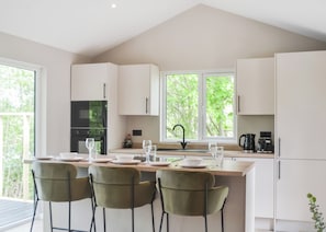 A modern kitchen with white cabinets, a central island with bar stools, and a view of greenery through the windows. - Buckingham Accessible (Doncaster, South Yorkshire)