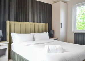 A modern hotel room with a large bed, a nightstand, a lamp, and a window with a view of greenery. - Buckingham Accessible (Doncaster, South Yorkshire)