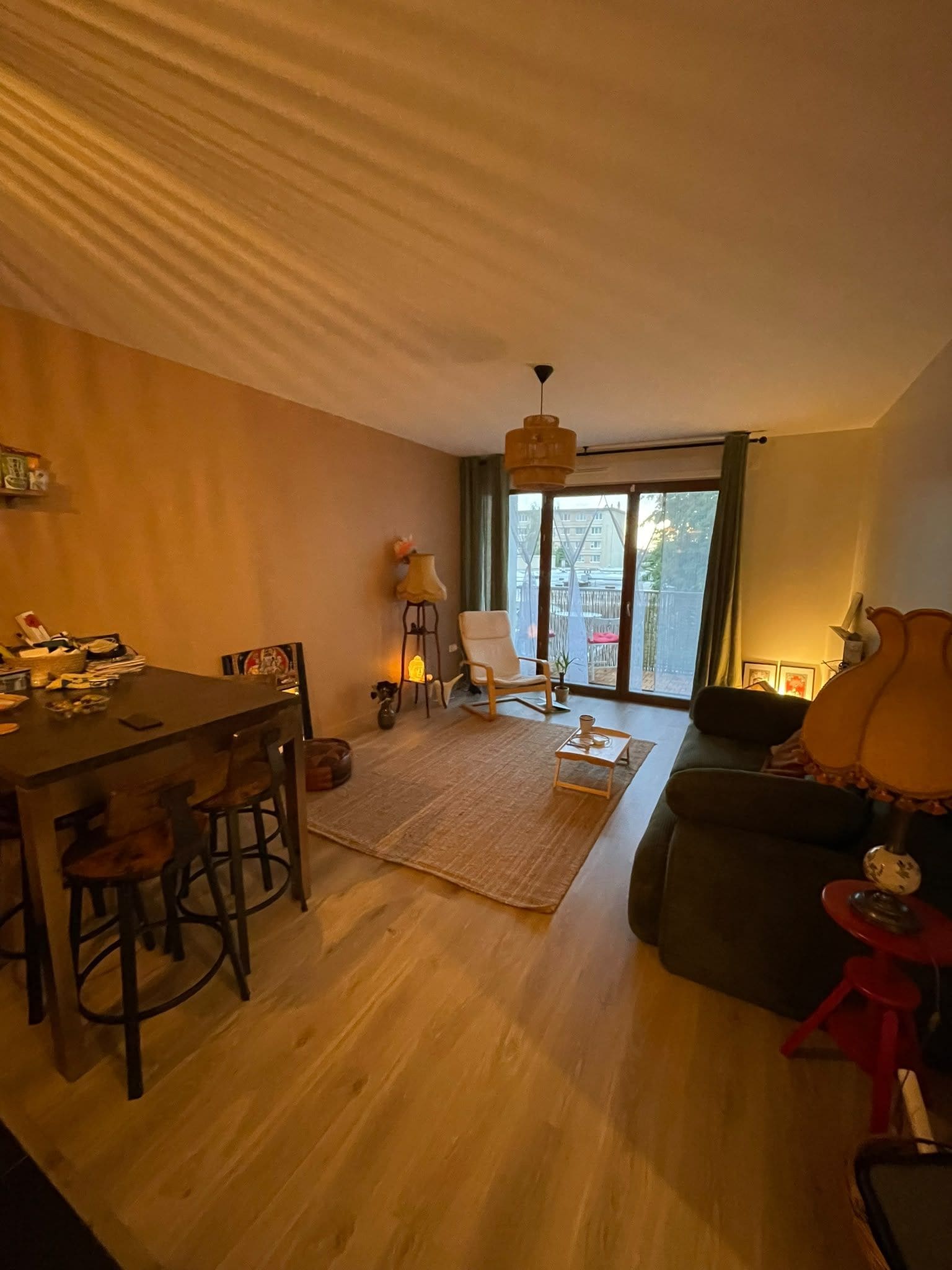 A living room with a dining area, a sofa, a red stool, a lamp, and a view through a glass door.