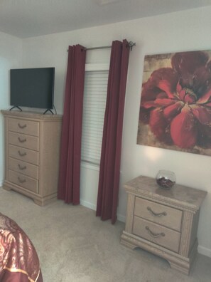 Een slaapkamer met een bed, een dressoir, een televisie erboven, een raam met gordijnen en een schilderij aan de muur.