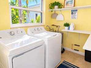 A laundry room with two white washers and dryers, a window with a view of palm trees, a notice board with laundry rules, and a shelf with a plant and a kettle. - Cozy 1BR Beach Retreat. Just Blocks from Clearwater Beach (Clearwater)
