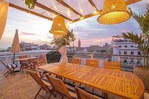 Uma área de refeições ao ar livre com vista para o horizonte da cidade, móveis de madeira e luminárias de vime suspensas.