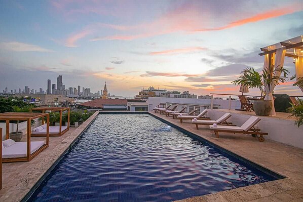 Piscina no terraço com espreguiçadeiras e vista para o horizonte da cidade ao pôr do sol.