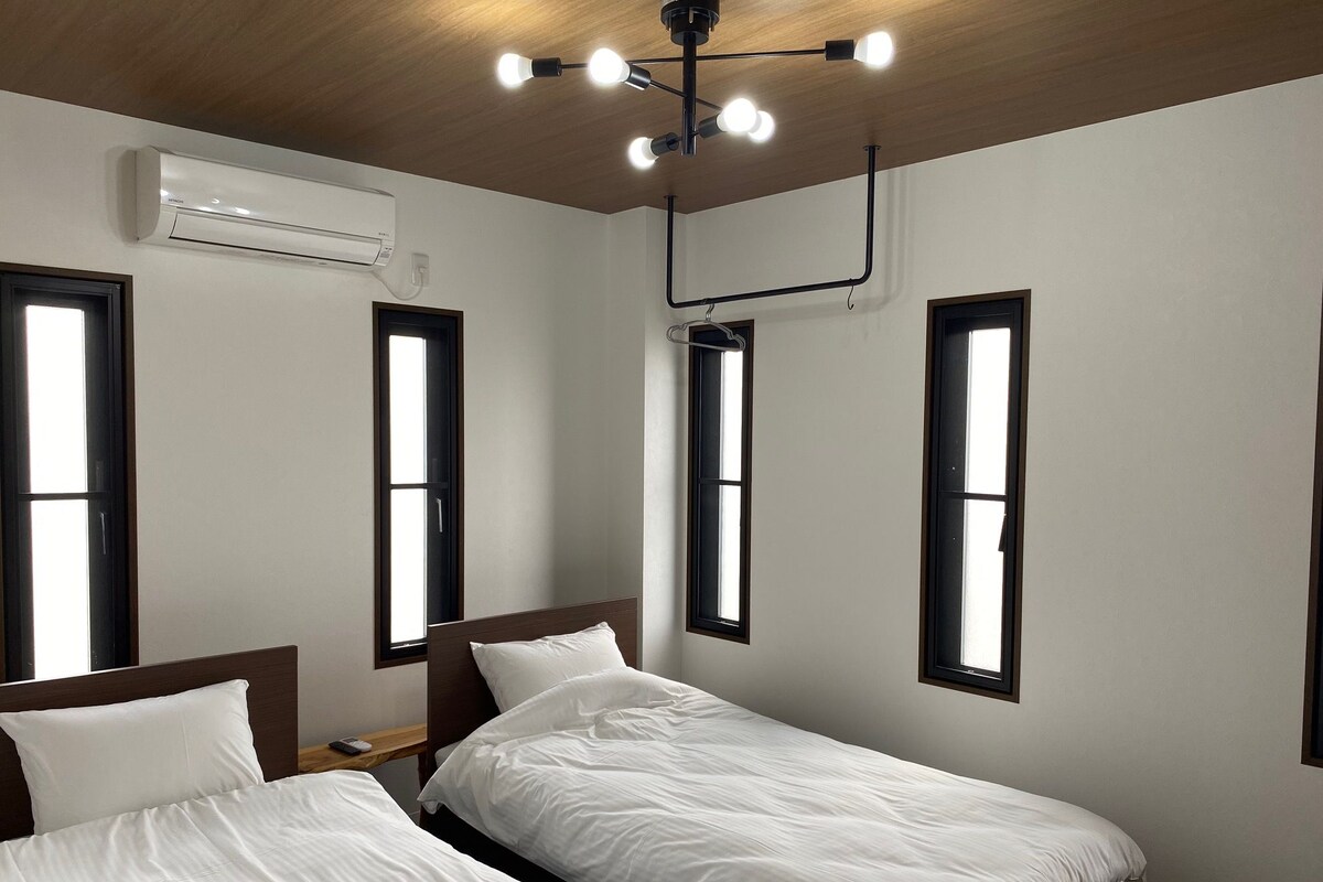 A hotel room with two beds, a ceiling fan, an air conditioning unit, and multiple windows.
