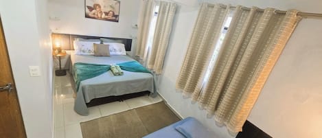 A hotel room with a bed, a bedside table, a lamp, a window with curtains, and a wall-mounted picture.