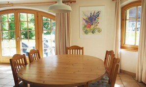 A dining area with a round wooden table, six chairs, a hanging light fixture, and a window with curtains. - BRIGHT 2-BEDROOM CHALET-STYLE APARTMENT WITH PRIVATE GARDEN ACCESS (Zermatt)