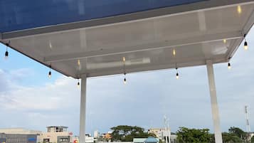 Un espacio al aire libre cubierto con techo metálico y luces colgantes, con vistas a una calle con edificios y una gasolinera.