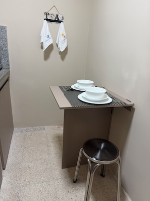 A kitchenette with a countertop, a small table with two white bowls and a spoon, a stool, and two towels hanging on a rack.