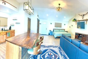 A spacious living area with a large wooden dining table, a blue sofa, and a bed with a green bedspread.
