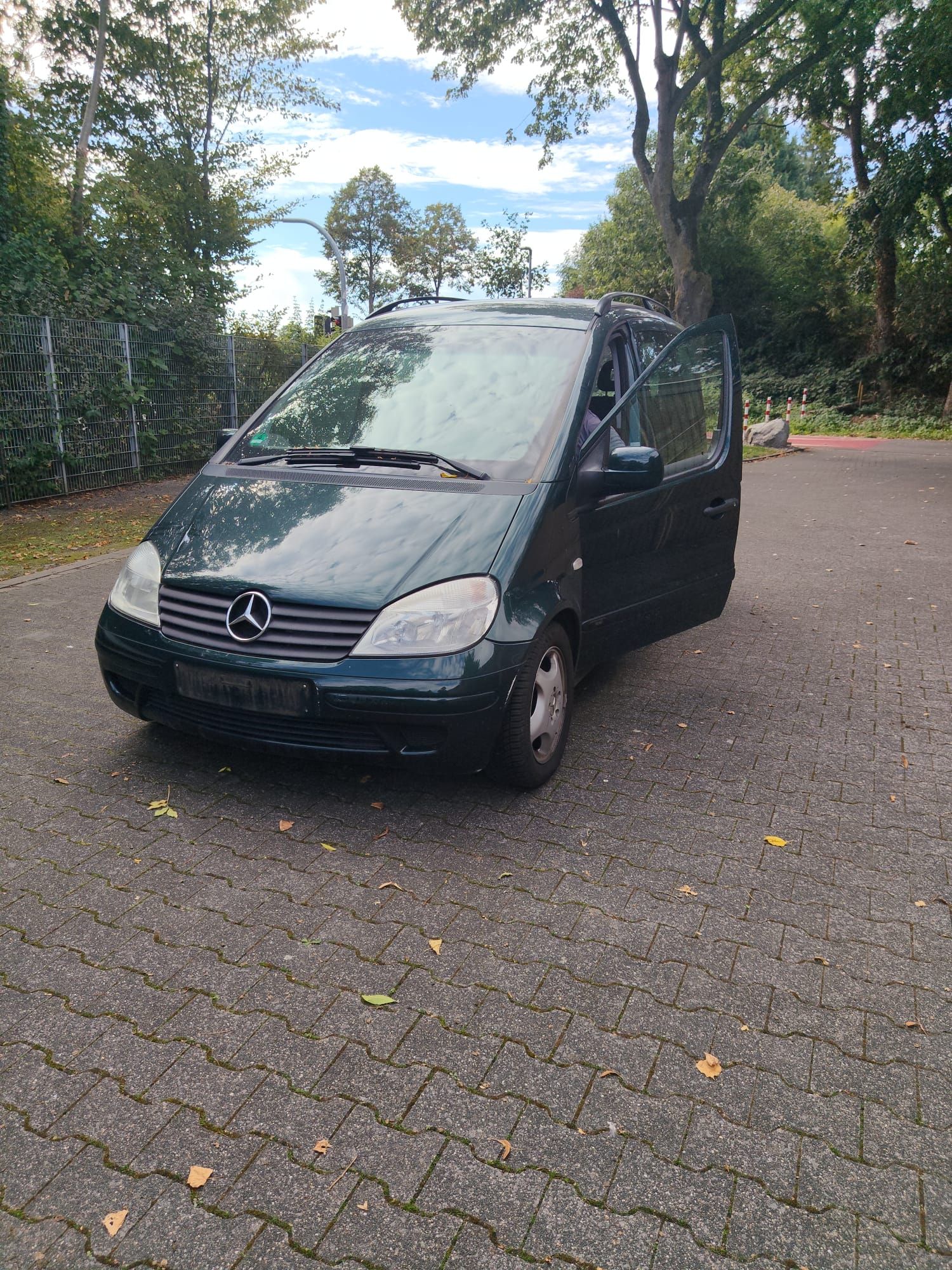 Ein geparktes Auto auf befestigter Fläche, im Hintergrund Bäume und ein Zaun.