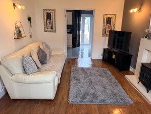 Ein Wohnzimmer mit einem weißen Ledersofa, einem grauen Teppich, Holzböden, einem Fernseher und einer Küche im Hintergrund.