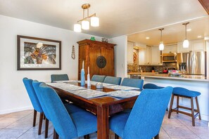 A dining area with a wooden table, blue upholstered chairs, and a framed picture on the wall. - Family-Friendly 4-Bedroom Retreat – Steps to the Pool & Across from the Ocean! (Kihei)