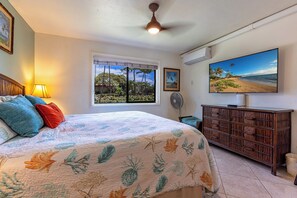 A bedroom with a bed, a window with a view of palm trees, a ceiling fan, and a TV on the wall. - Family-Friendly 4-Bedroom Retreat – Steps to the Pool & Across from the Ocean! (Kihei)