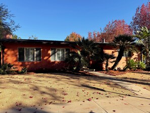 A single-story house with a red exterior, a front yard with scattered leaves, and a clear blue sky.
