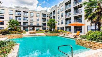 A swimming pool with lounge chairs and umbrellas, surrounded by a multi-story building with balconies.