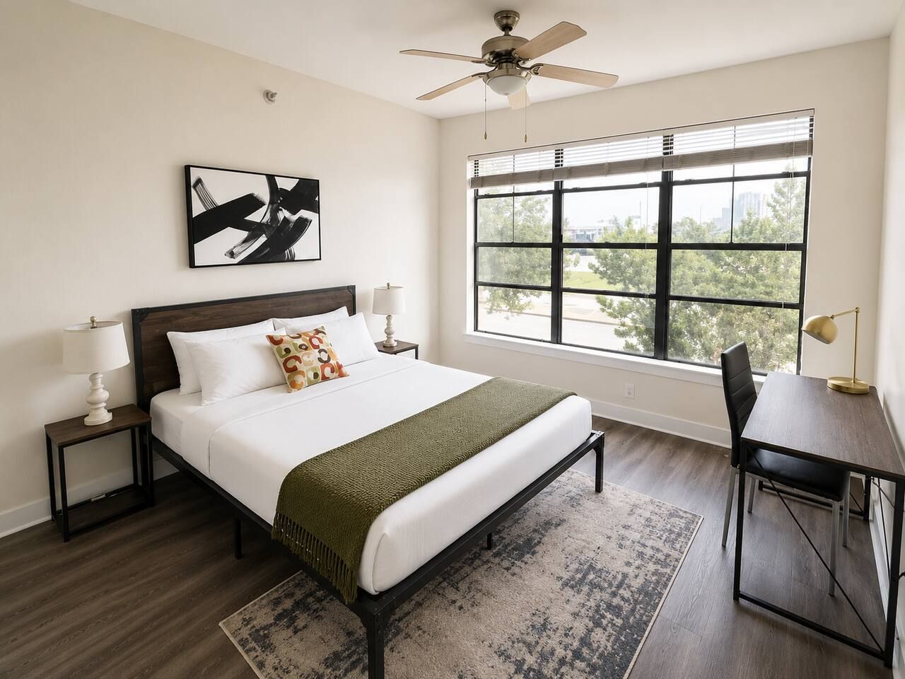Un dormitorio con cama, mesitas de noche, escritorio, silla, ventana con persianas, ventilador de techo y un cuadro en la pared.