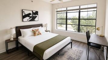 Un dormitorio con cama, mesitas de noche, escritorio, silla, ventana con persianas, ventilador de techo y un cuadro en la pared.