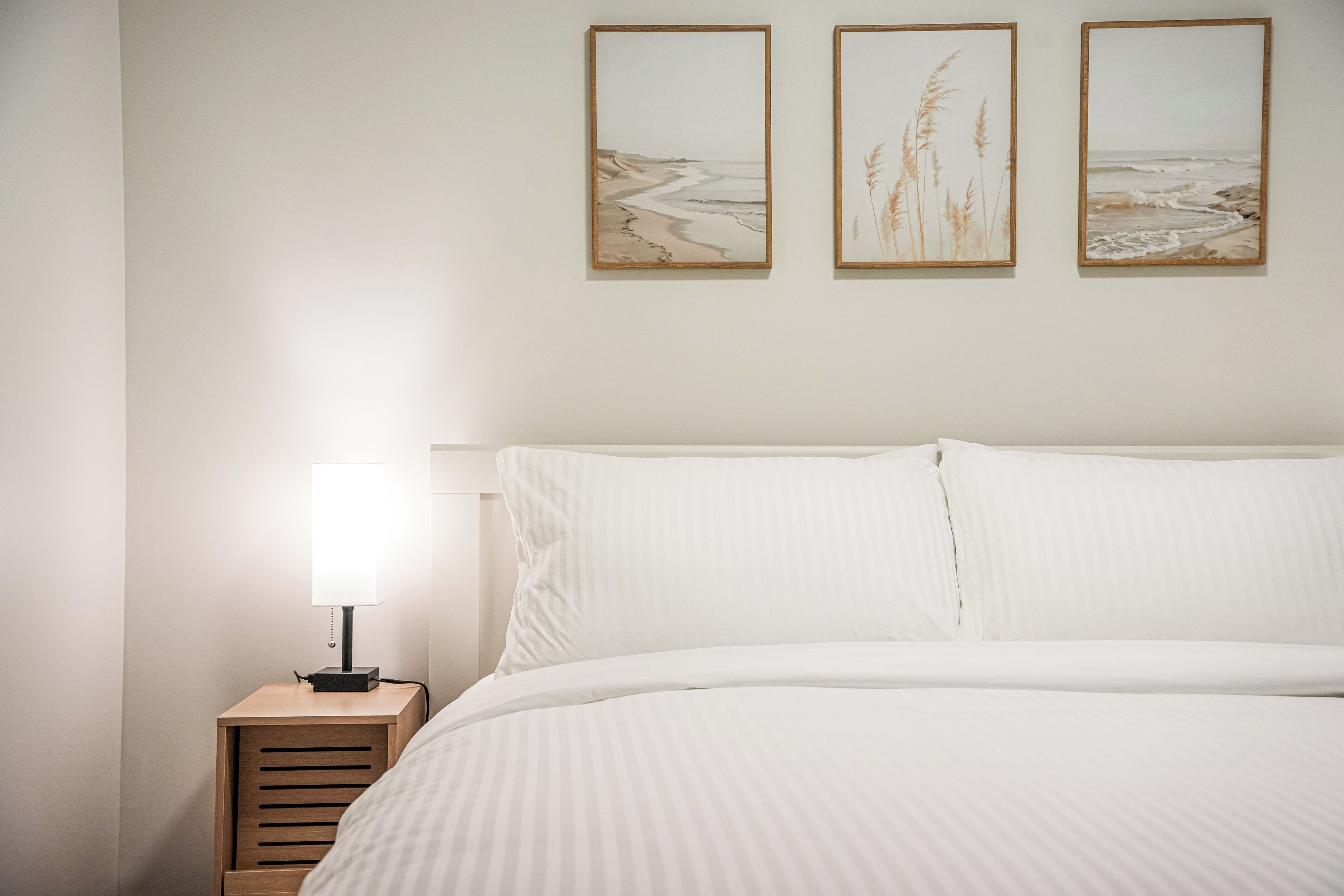 A neatly made bed with white linens, a bedside table with a lamp, and three framed artworks on the wall.