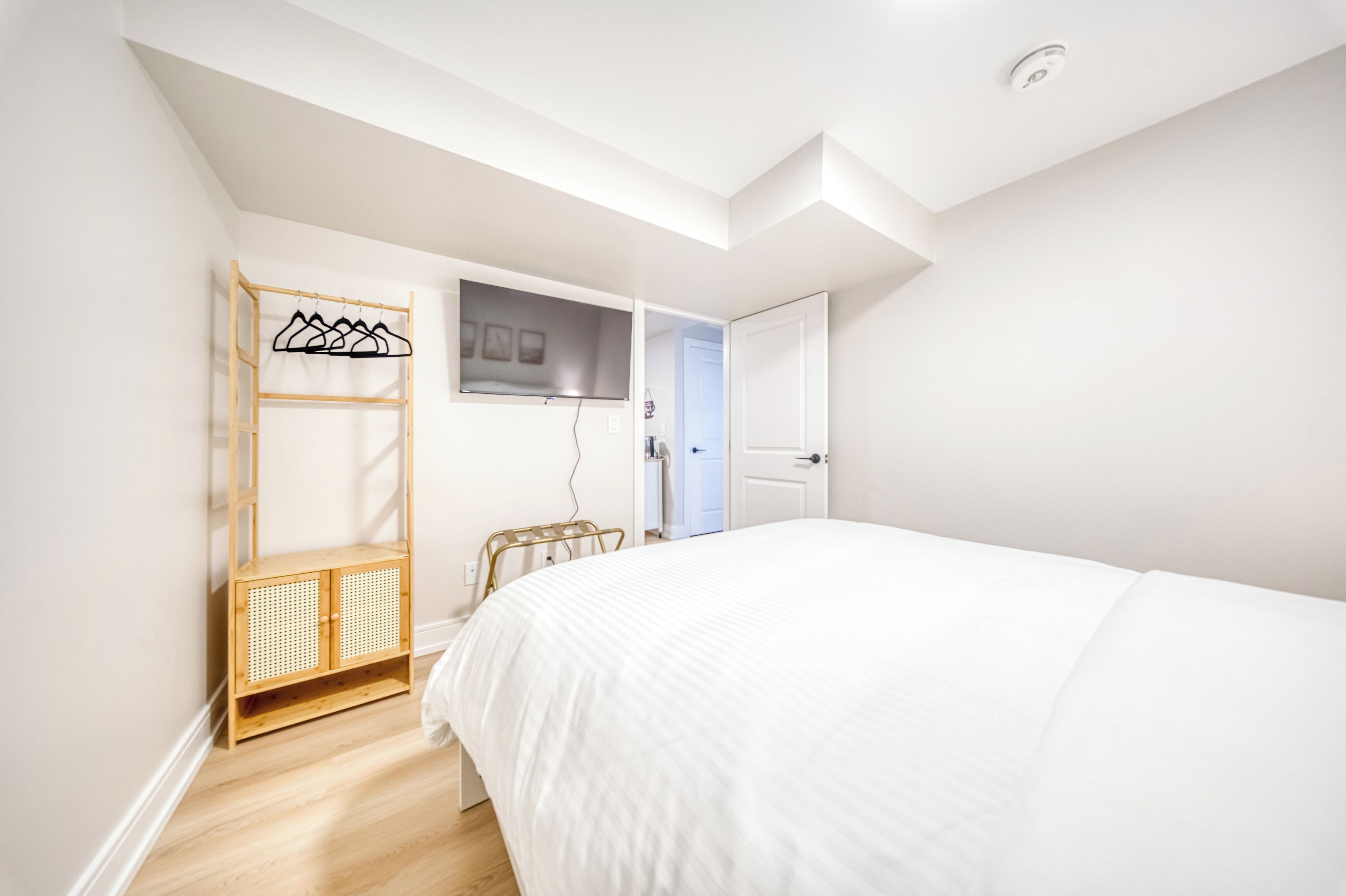 A bedroom with a bed, a wooden wardrobe, a television mounted on the wall, and a door leading to another room.