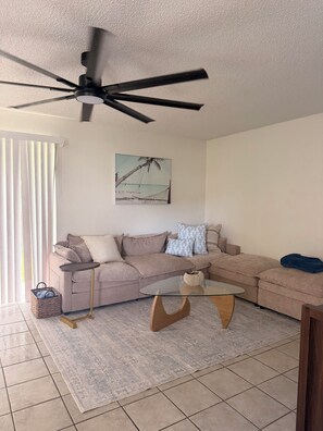 A living room with a beige sectional sofa, a glass coffee table, a ceiling fan, and a beach-themed wall art. - Welcome to the Sandy Beach Oasis, your relaxing New Smyrna Beach getaway! (New Smyrna Beach)