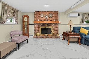 A living room with a brick fireplace, a clock, a sofa, a bench, and a lamp. - Spacious 4BR Home Near Binghamton & UHS (Endicott)