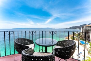 A balcony with a glass table and two wicker chairs, overlooking a beach and a hotel building. - Cozy oceanfront studio (Puerto Vallarta)