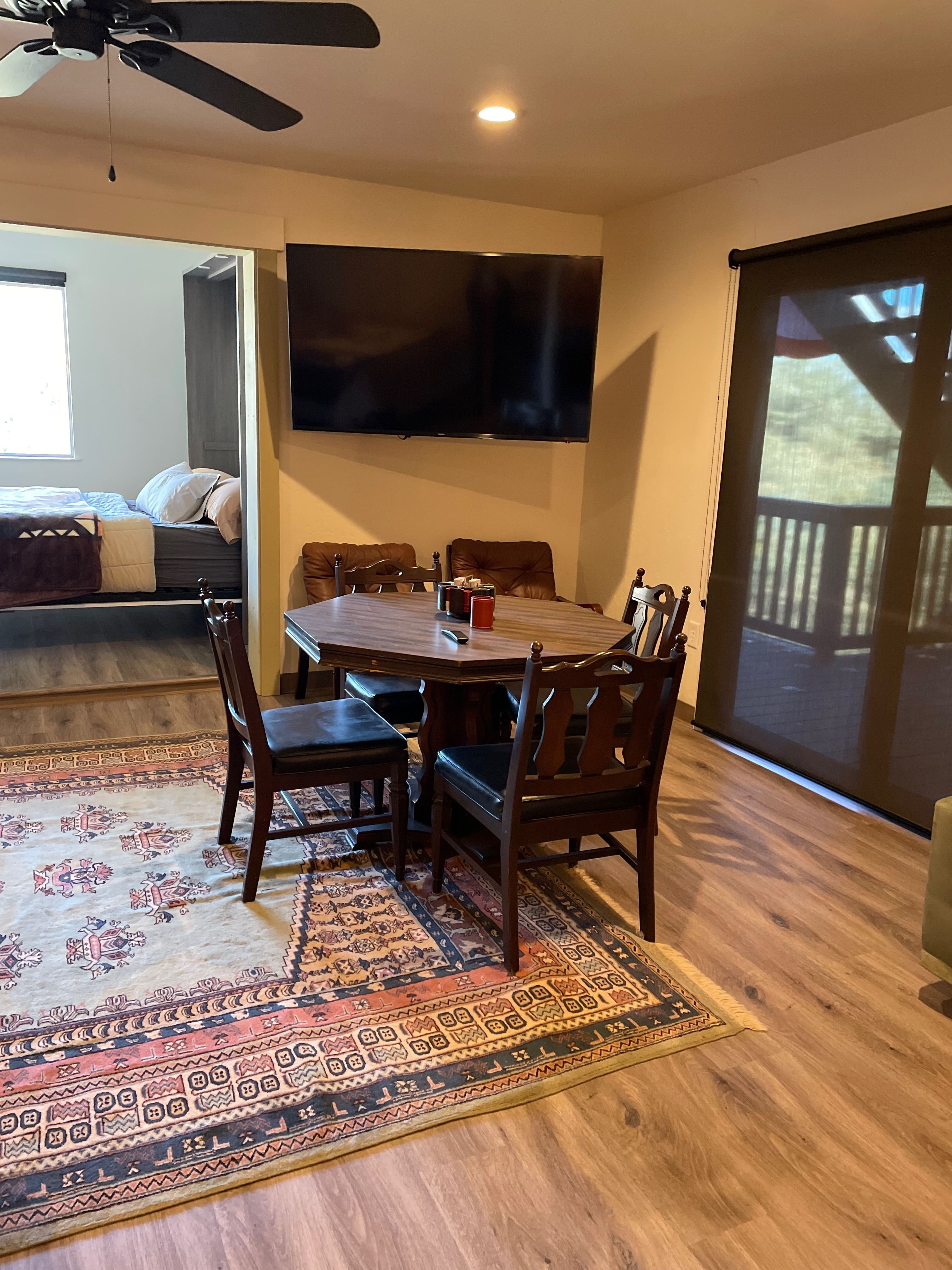 Un espace repas comprenant une grande table, des chaises, un téléviseur fixé au mur et une vue sur l’extérieur grâce à une porte coulissante.