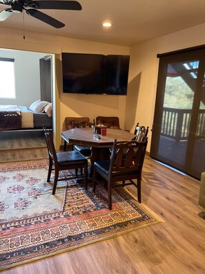 Un espace repas comprenant une grande table, des chaises, un téléviseur fixé au mur et une vue sur l’extérieur grâce à une porte coulissante.
