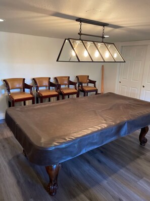 A rectangular pool table with a dark felt surface, surrounded by four wooden chairs with cushioned seats. A modern, geometric light fixture hangs above the table. - Beautiful Large Wood Cabin with Large Covered Deck (Bradley)