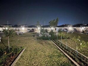 Ein Parkplatz mit mehreren Autos und ein gut beleuchteter Gartenbereich mit Wegen und kleinen Bäumen.