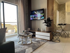 Ein modernes Wohnzimmer mit einem an der Wand befestigten Flachbildfernseher, einem Essbereich mit Tisch und Stühlen und einem großen Fenster mit Blick nach draußen.